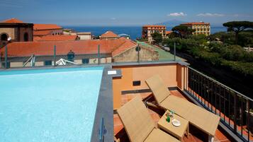 Una piscina en la azotea con sillones de descanso y una mesa, con vistas a un paisaje urbano con edificios de tejados rojos y vistas al mar.
