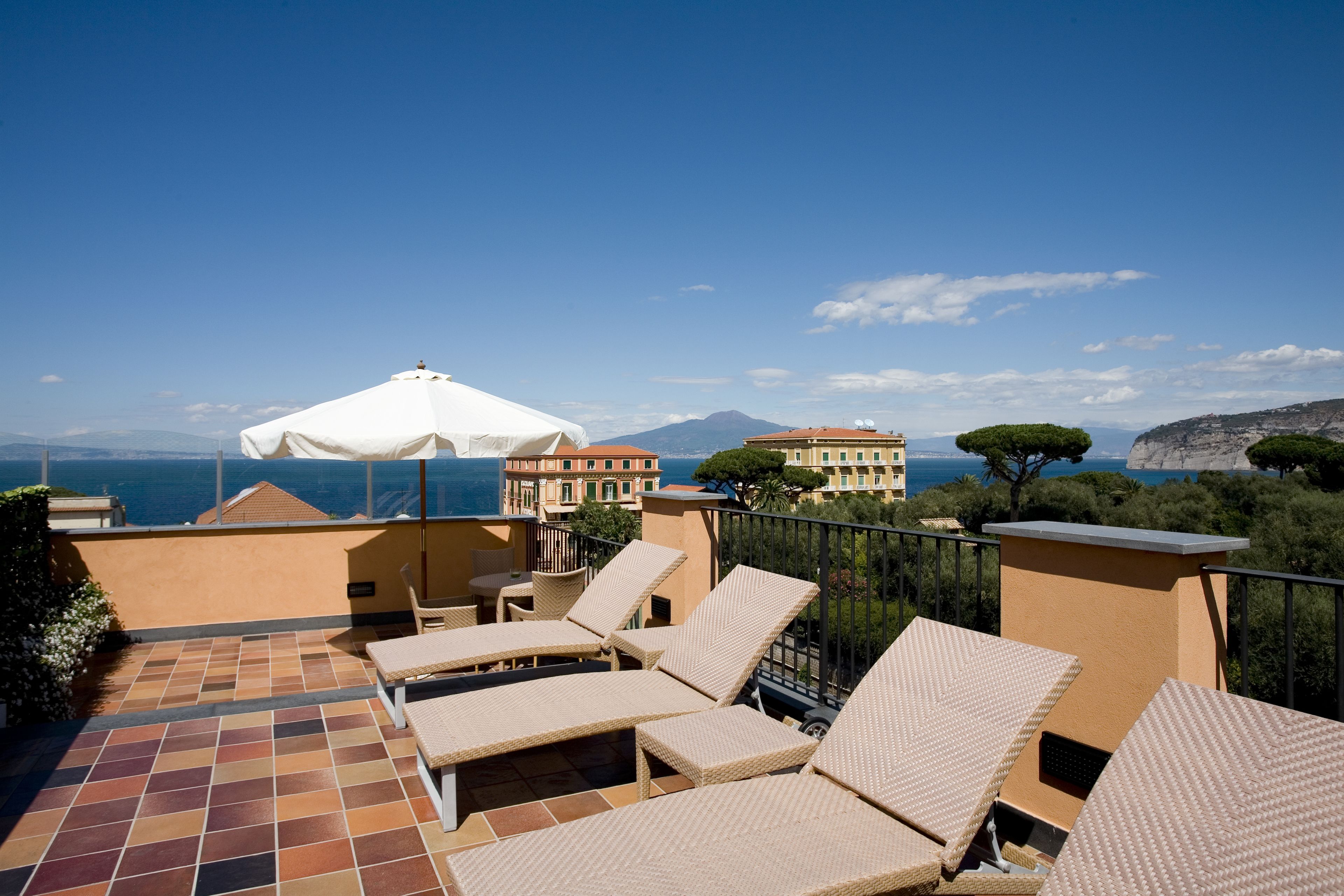 A terrace with lounge chairs and a white umbrella, overlooking a coastal view with buildings and a mountain in the distance.