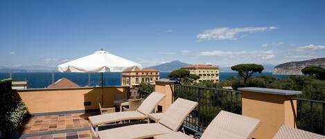 A terrace with lounge chairs and a white umbrella, overlooking a coastal view with buildings and a mountain in the distance.