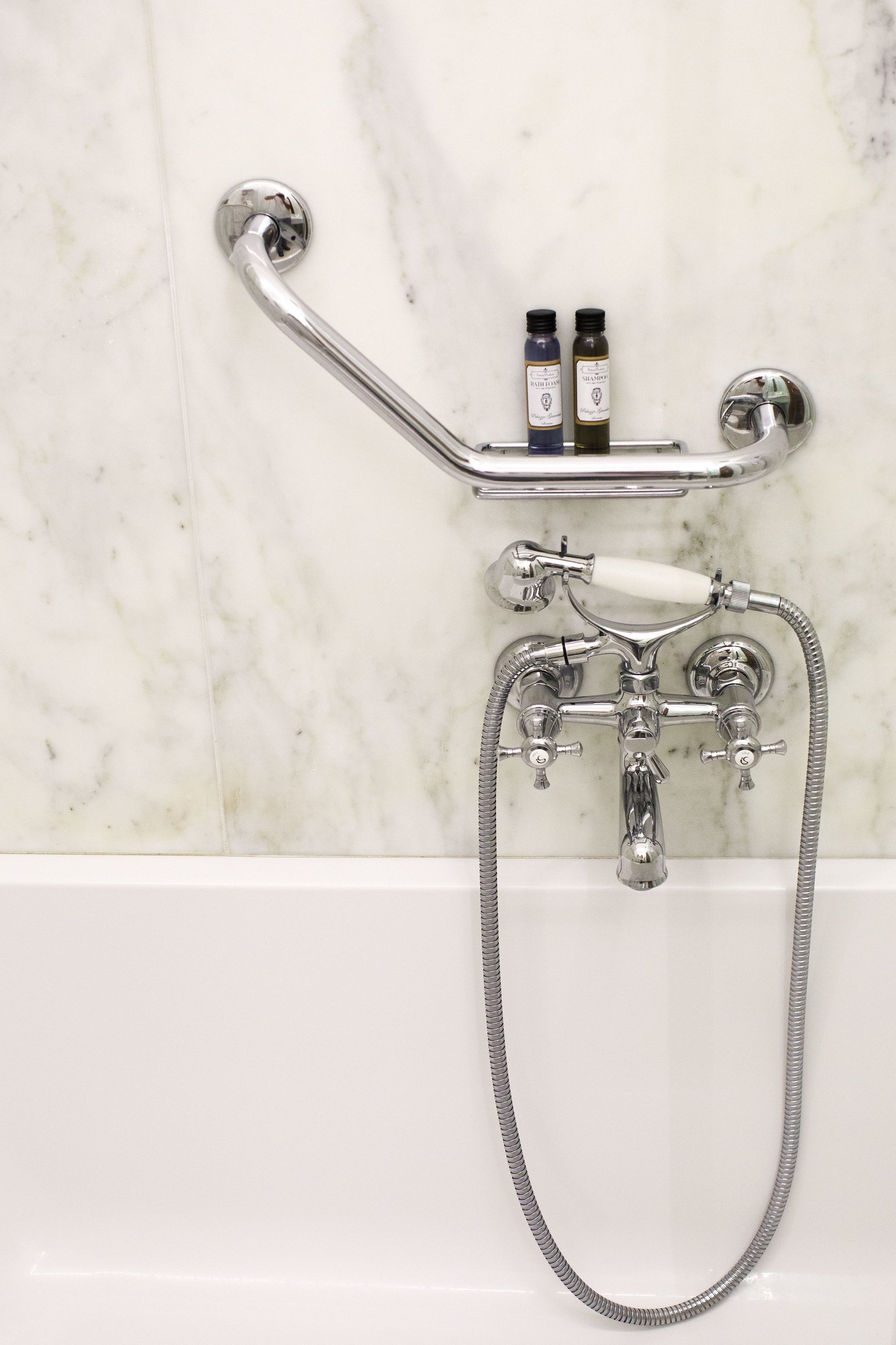 Un baño con pared de mármol blanco, ducha cromada con cabezal de ducha de mano y dos botellas de champú en un estante cromado.