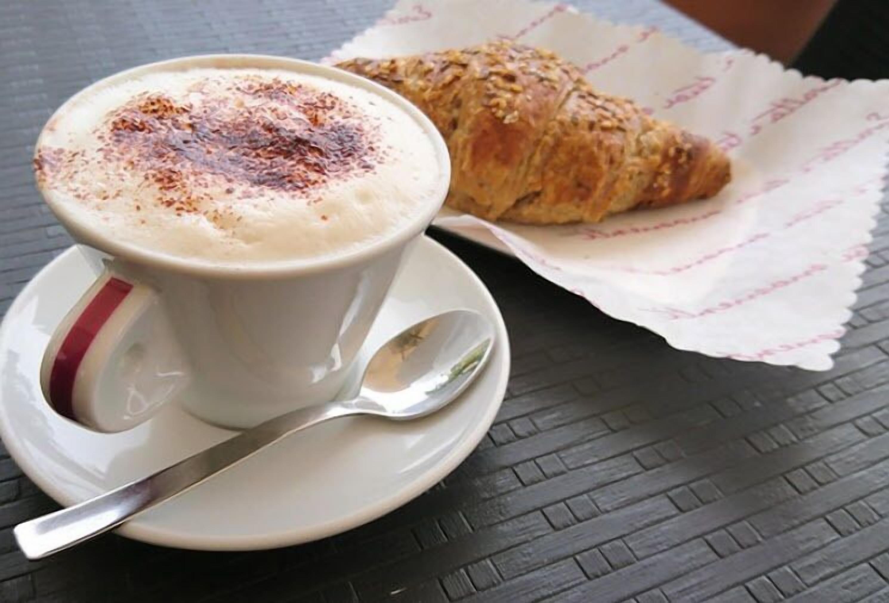 A cup of coffee with a frothy top and a croissant on a table.
