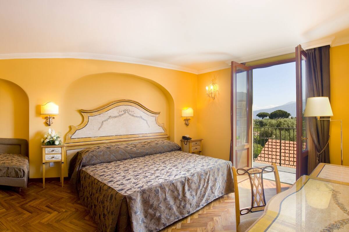 Habitación de hotel con cama, una silla, una mesita de noche, una lámpara y vistas al exterior.