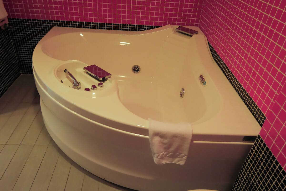 A large bathtub with a faucet and a small shelf, surrounded by pink and black tiles.
