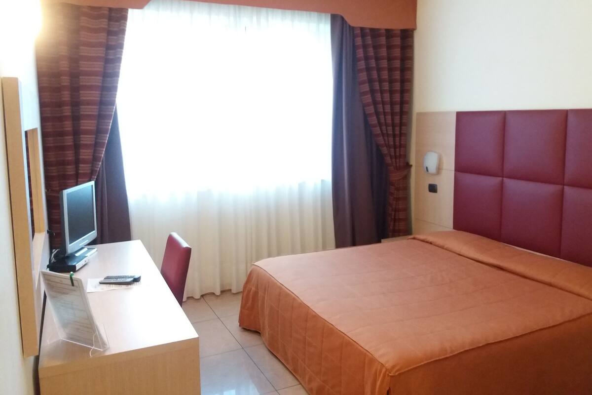 A hotel room with a bed, a desk with a computer, a chair, and a window with curtains.