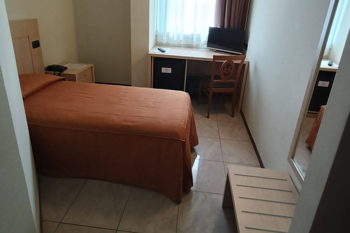 A hotel room with a bed, a desk with a computer, a chair, a small table, and a mirror.