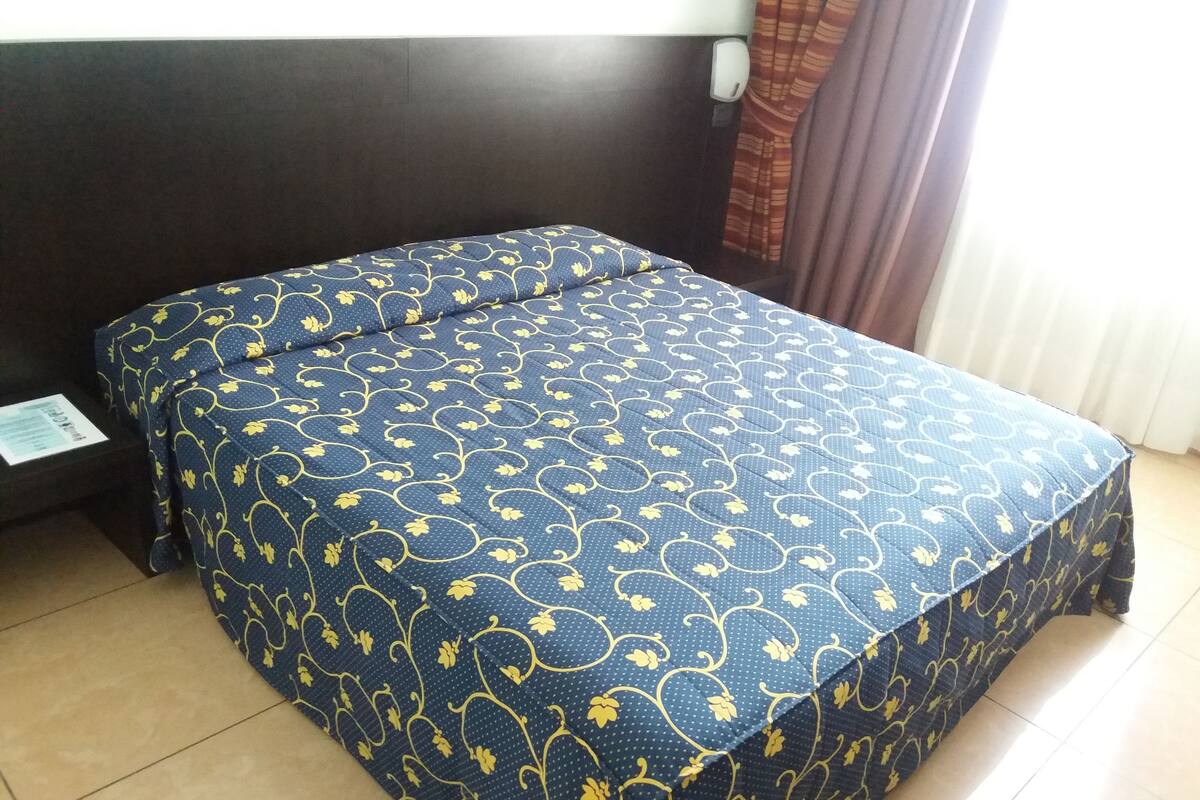 A neatly made bed with a floral pattern, a dark headboard, and a window with curtains.