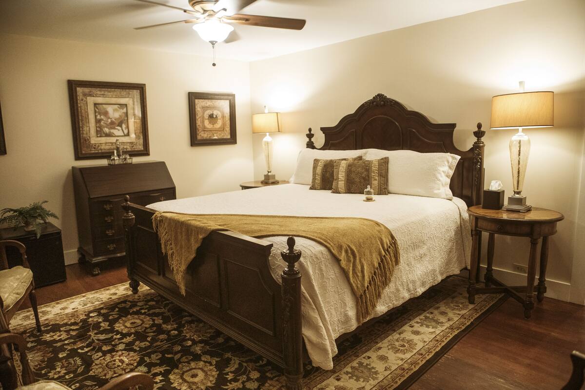 A traditional bedroom with a wooden bed, two bedside tables with lamps, a ceiling fan, and a fireplace.