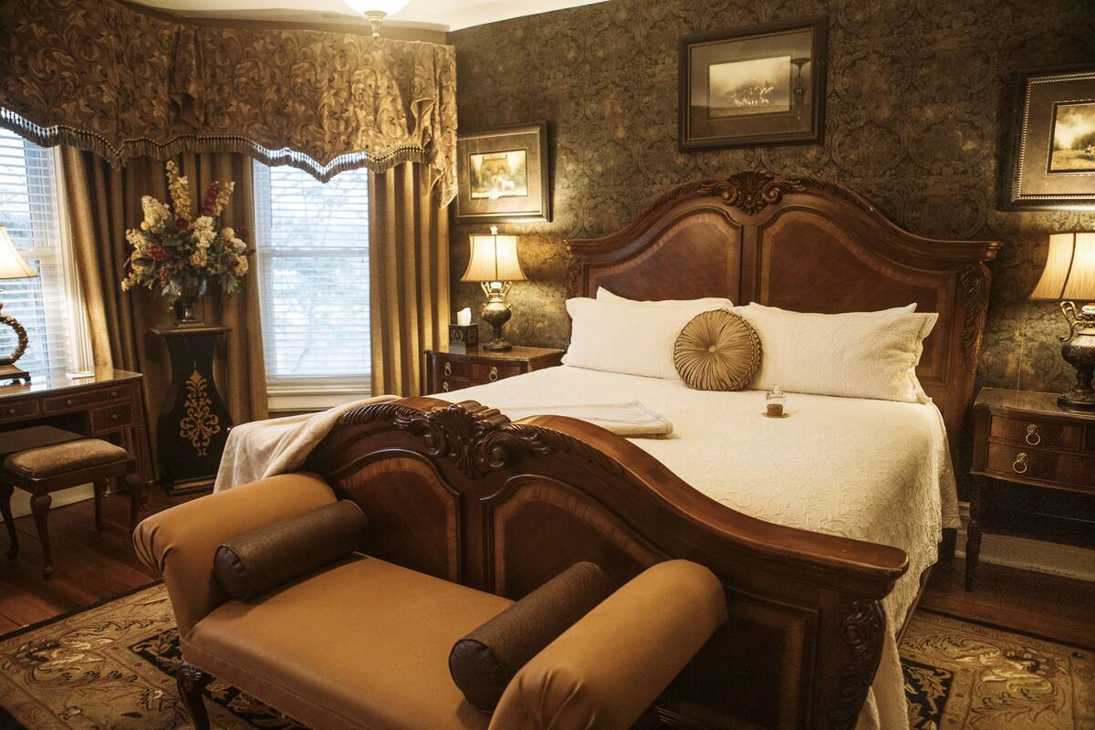 A classic hotel room with a large bed, a wooden headboard, a sitting area with a chair, a desk, and a nightstand with a lamp.