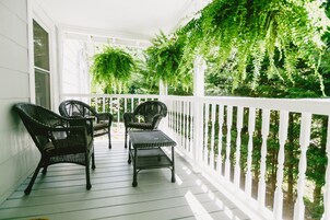 Une véranda couverte, meublée de meubles en osier et dotée d’une balustrade blanche, entourée d’une végétation luxuriante.
