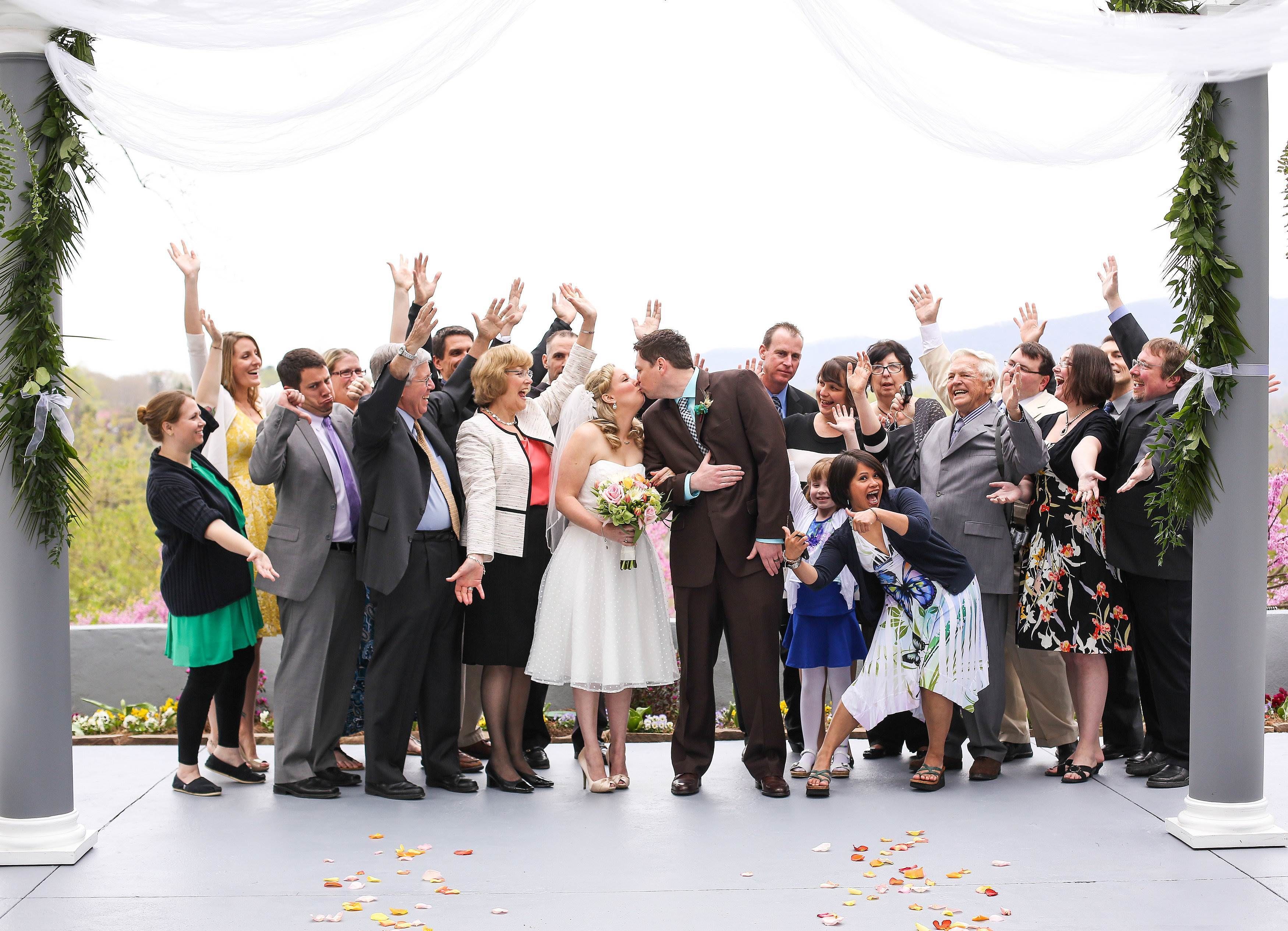 A group of people gathered under a white canopy with greenery on either side, standing on a tiled floor with scattered flower petals.