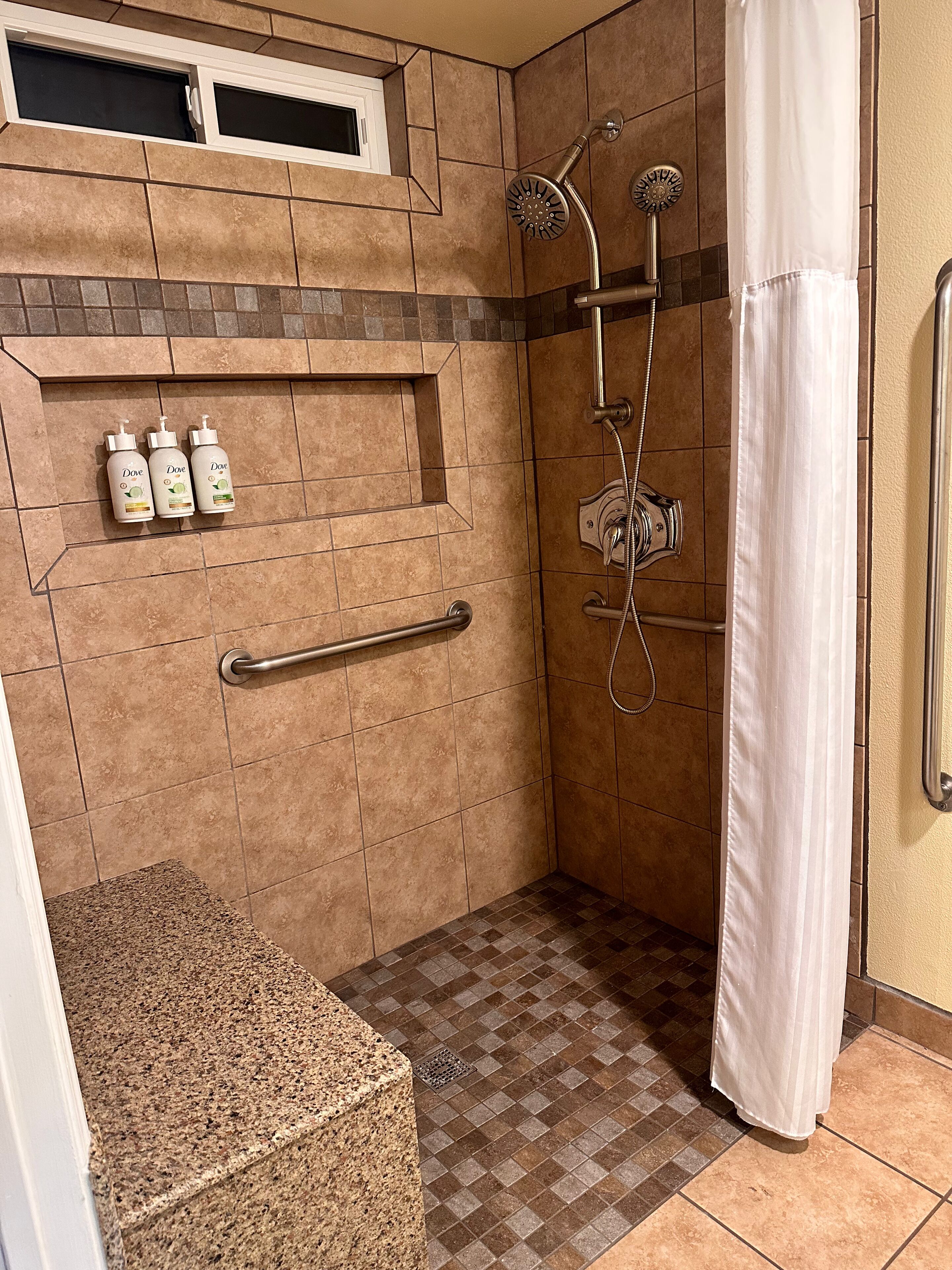 A shower area with a grab bar, a showerhead, and three Dove shampoo bottles on a shelf.