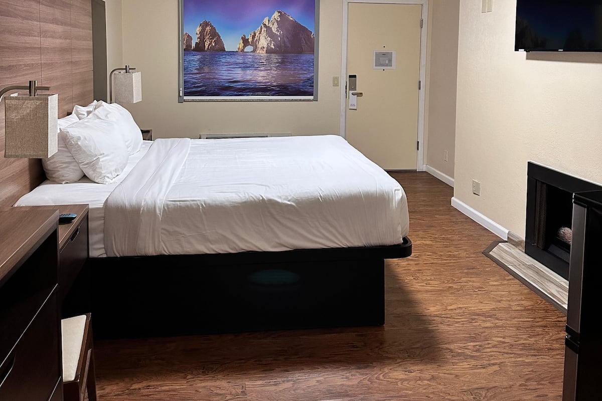 A hotel room with a large bed, a flat-screen TV mounted on the wall, a painting of a mountainous landscape, and a fireplace.