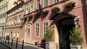 Un bâtiment rose avec de nombreuses fenêtres et une porte noire. Des jardinières sont posées sur les rebords des fenêtres et un banc se trouve à l’extérieur.