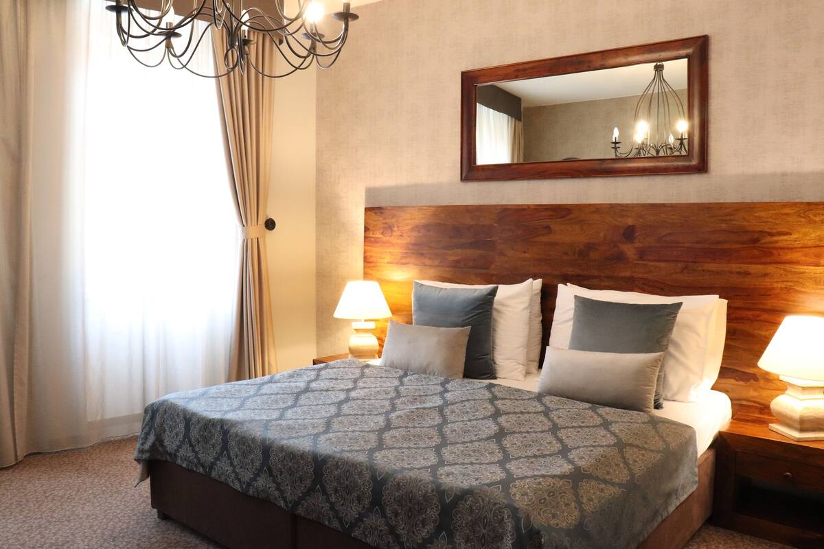 Une chambre d’hôtel avec une tête de lit en bois, un grand lit avec une literie à motifs, deux lampes de chevet et un miroir au-dessus du lit.