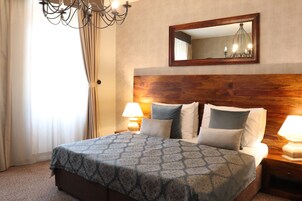 Quarto de hotel com cabeceira de madeira, uma cama grande com roupa de cama estampada, duas luminárias de cabeceira e um espelho sobre a cama.