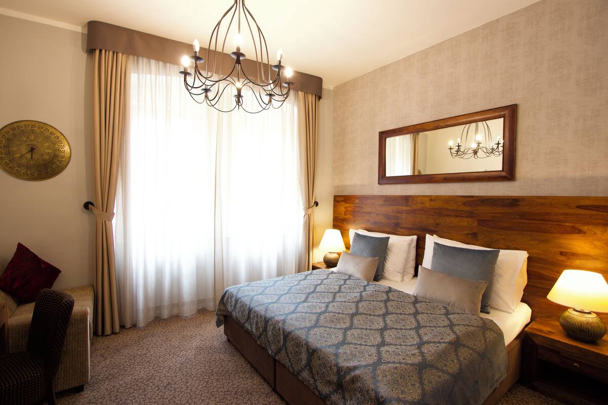Une chambre d’hôtel avec un grand lit, un lustre, une tête de lit en bois, une table de chevet avec une lampe, une chaise et une horloge murale.