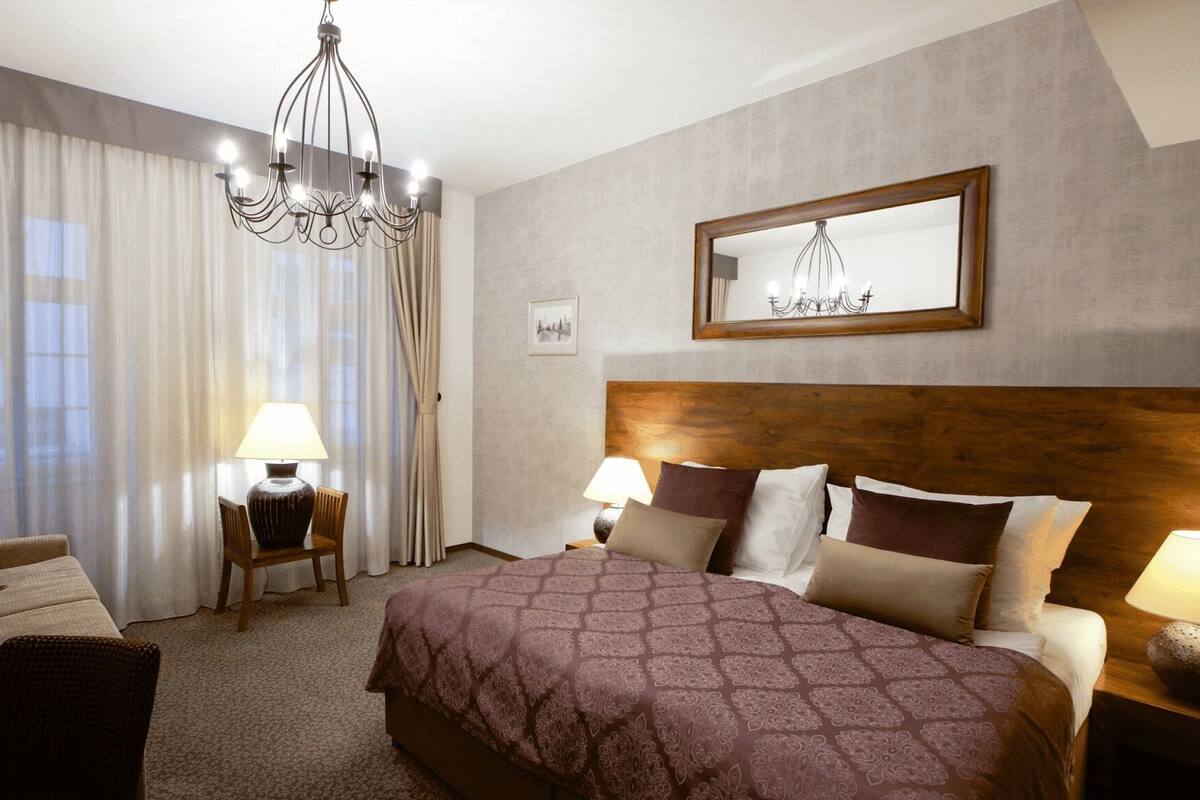 Une chambre d’hôtel avec un grand lit, deux tables de chevet avec des lampes, une chaise, un lustre et un tableau encadré au mur.