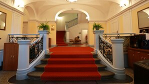 Eine imposante Treppe mit rotem Teppich, der zu einer zweiten Etage führt, gesäumt von weißen Säulen und schwarzen Eisengeländern.