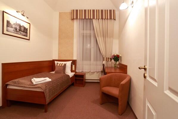 Quarto de hotel com uma cama de casal, uma poltrona, uma mesinha com um vaso e uma janela com cortinas leves.