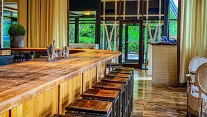 A bar area with a long wooden counter, multiple stools, and hanging pendant lights. There are large windows with a view of greenery outside. - Proximity Hotel (Greensboro)