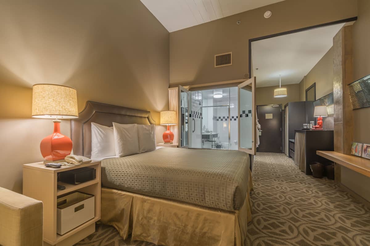 A hotel room with a bed, bedside lamps, a nightstand, a desk, and a bathroom visible through an open door.