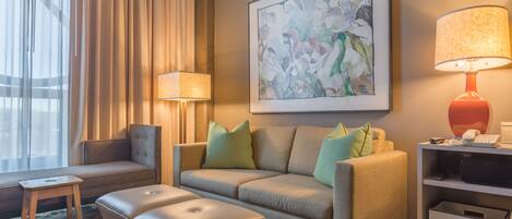 Una camera d'albergo moderna con un divano, poggiapiedi, tavolino, lampada e un'opera d'arte appesa al muro.