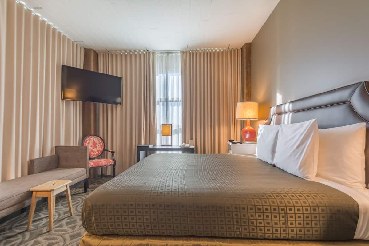 A hotel room with a large bed, a flat-screen TV mounted on the wall, a sofa, a small table, and a nightstand with a lamp.