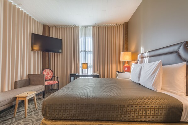 A hotel room with a large bed, a flat-screen TV mounted on the wall, a sofa, a small table, and a nightstand with a lamp.