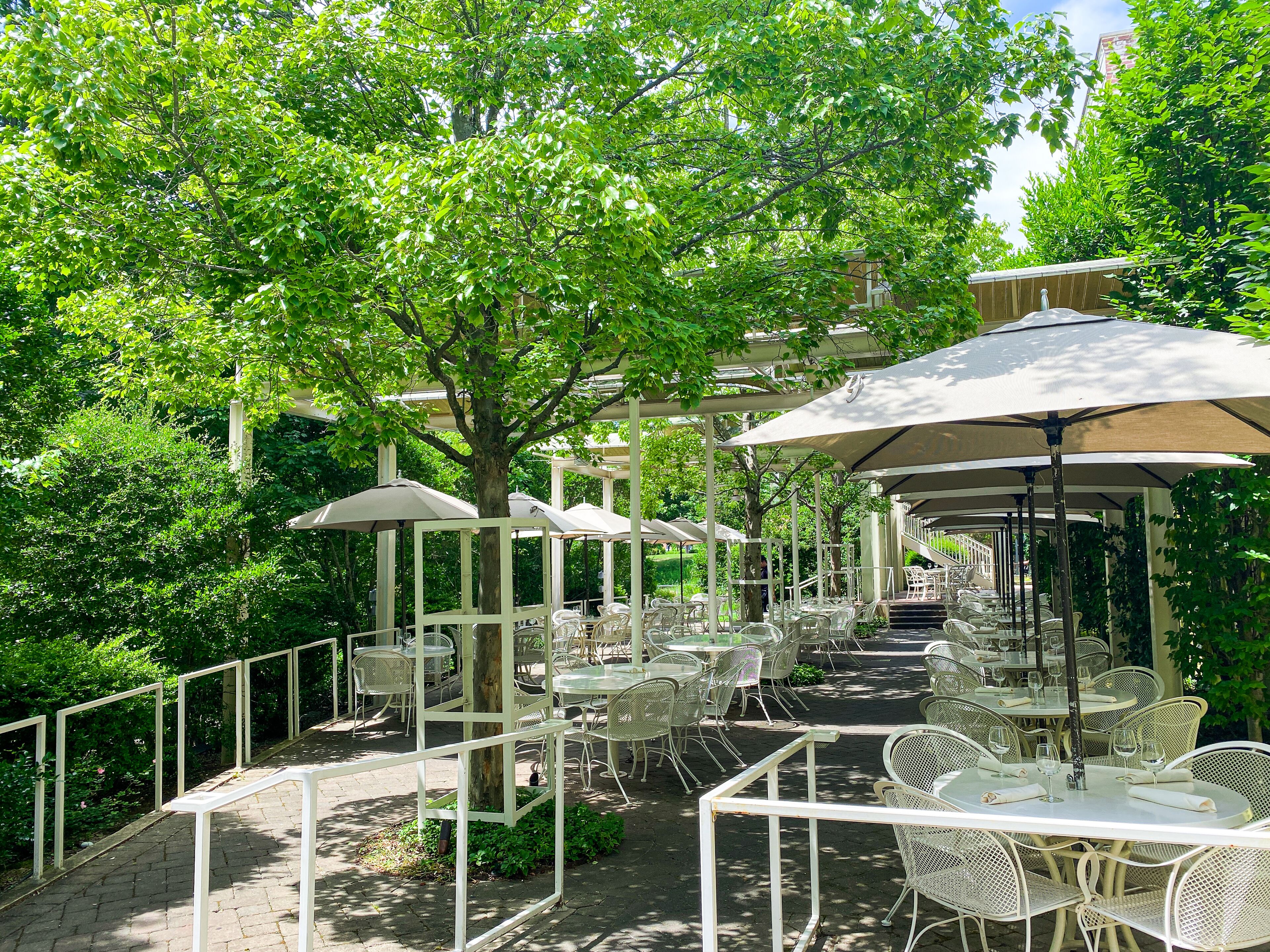 Eine Terrasse im Freien mit weißen Metalltischen und -stühlen, umgeben von Bäumen und beschattet durch große Sonnenschirme.