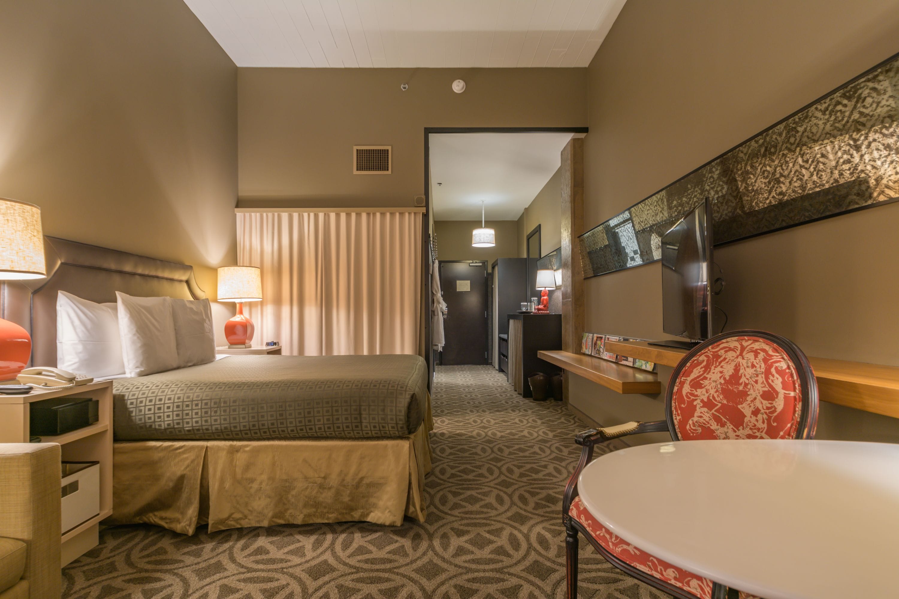 Habitación de hotel con cama, mesita de noche, lámpara, silla y escritorio con televisor.