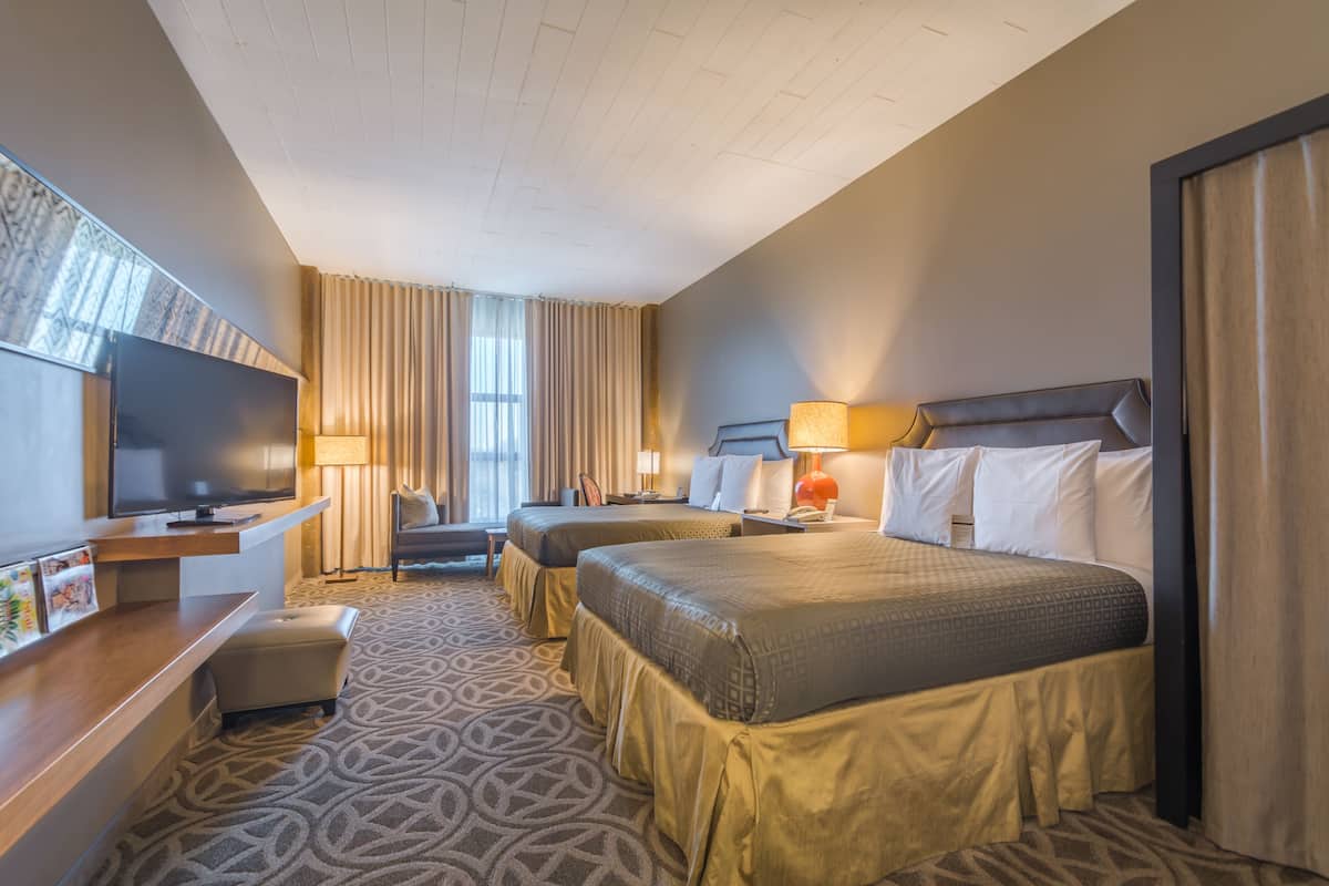 A hotel room with two beds, a desk with a flat-screen TV, a chair, and a large window with curtains.