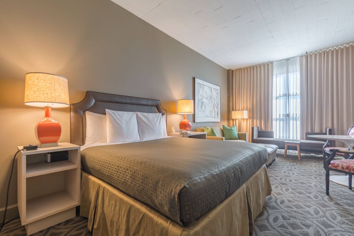 A hotel room with a large bed, two bedside tables with lamps, a seating area with a chair and a small table, and a large window with curtains.
