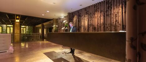 A reception area with a modern design, featuring a reception desk, a patterned wall, and a spacious lobby with a high ceiling and recessed lighting.