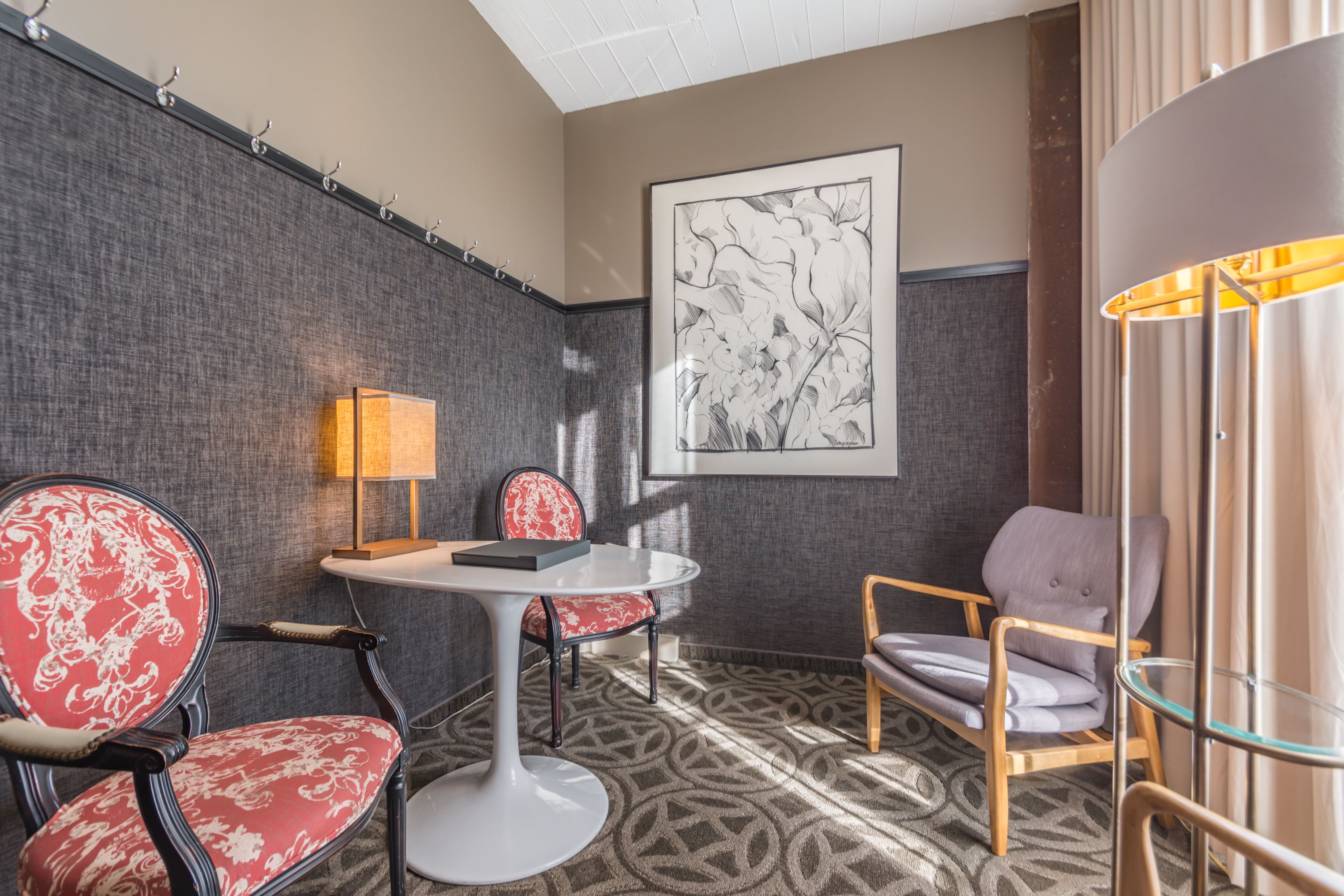 Une chambre d’hôtel avec un tapis à motifs, une table ronde blanche, deux chaises avec un revêtement rouge et gris, une grande œuvre d’art encadrée au mur et un lampadaire.