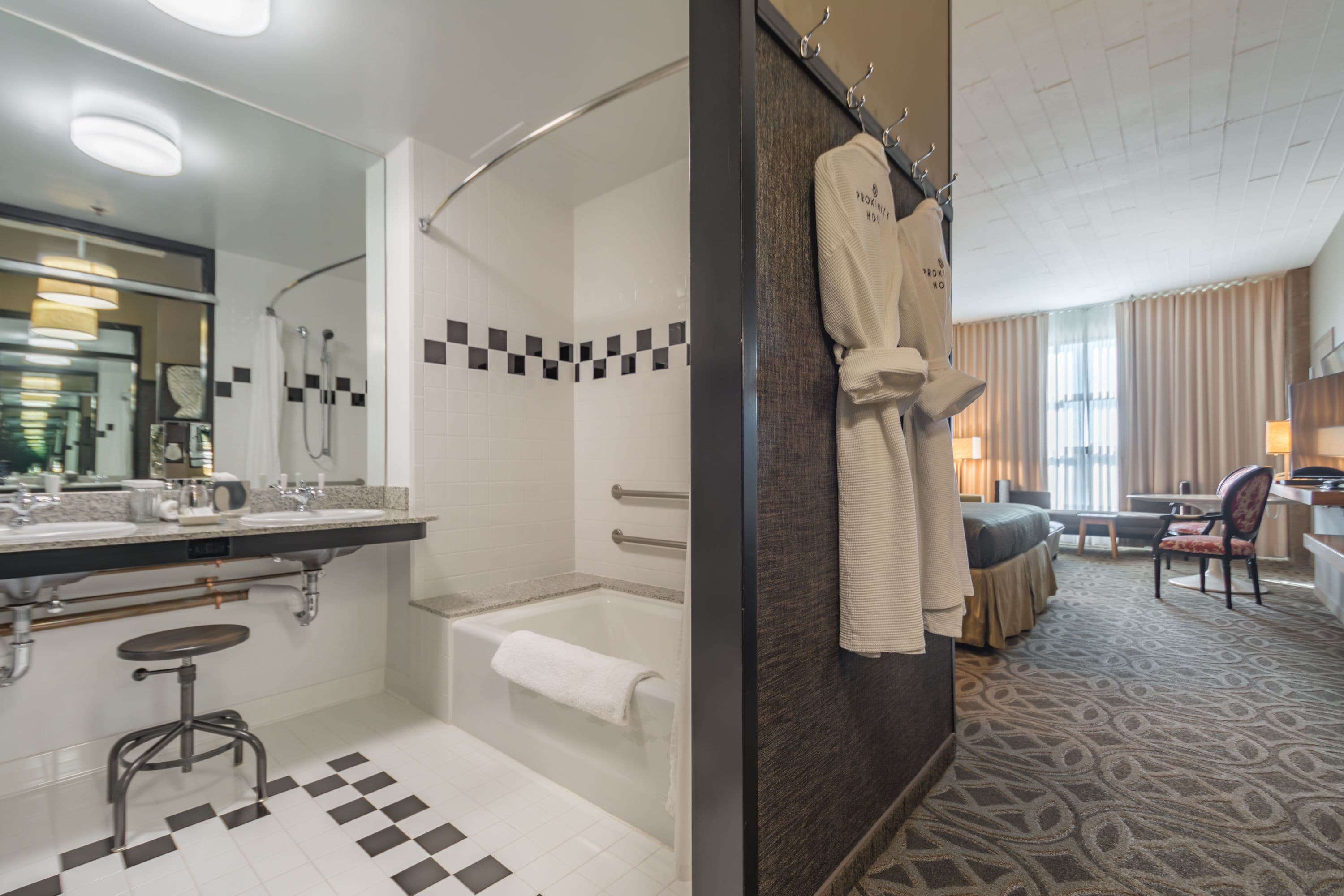 A hotel bathroom with a bathtub, a shower, a sink, and a mirror. There is a robe hanging on the wall and a chair in the corner.