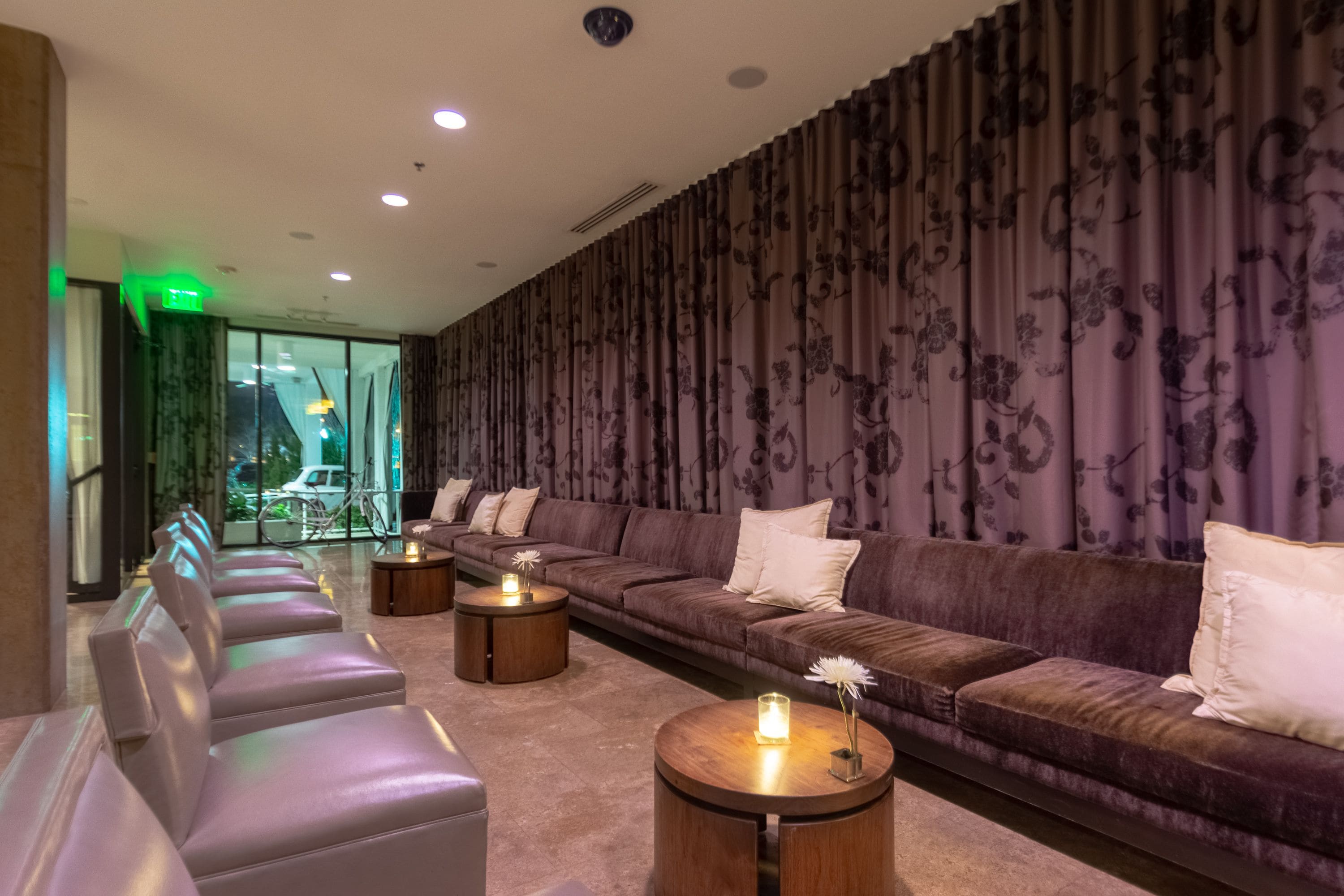 A modern lounge with a sofa, chairs, and round tables, featuring a large window and a patterned wall.