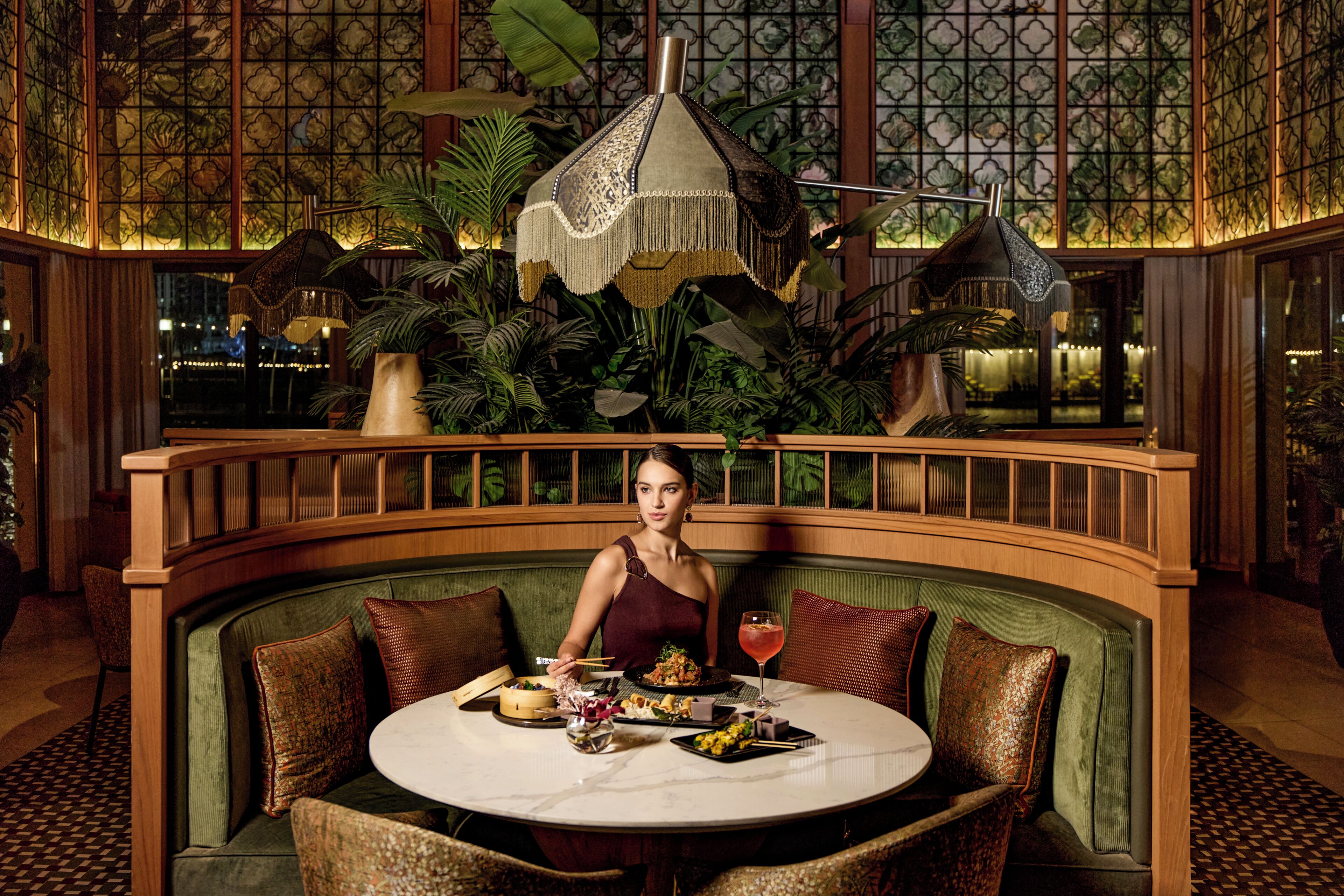 Une femme est assise à une table ronde, devant une variété de plats. La pièce est décorée d’un banc recouvert d’un tissu vert, de lambris en bois et d’un grand lampadaire suspendu.