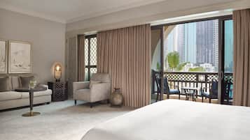 A hotel room with a large bed, a sofa, a chair, a small table with a lamp, and a view of tall buildings through the window.