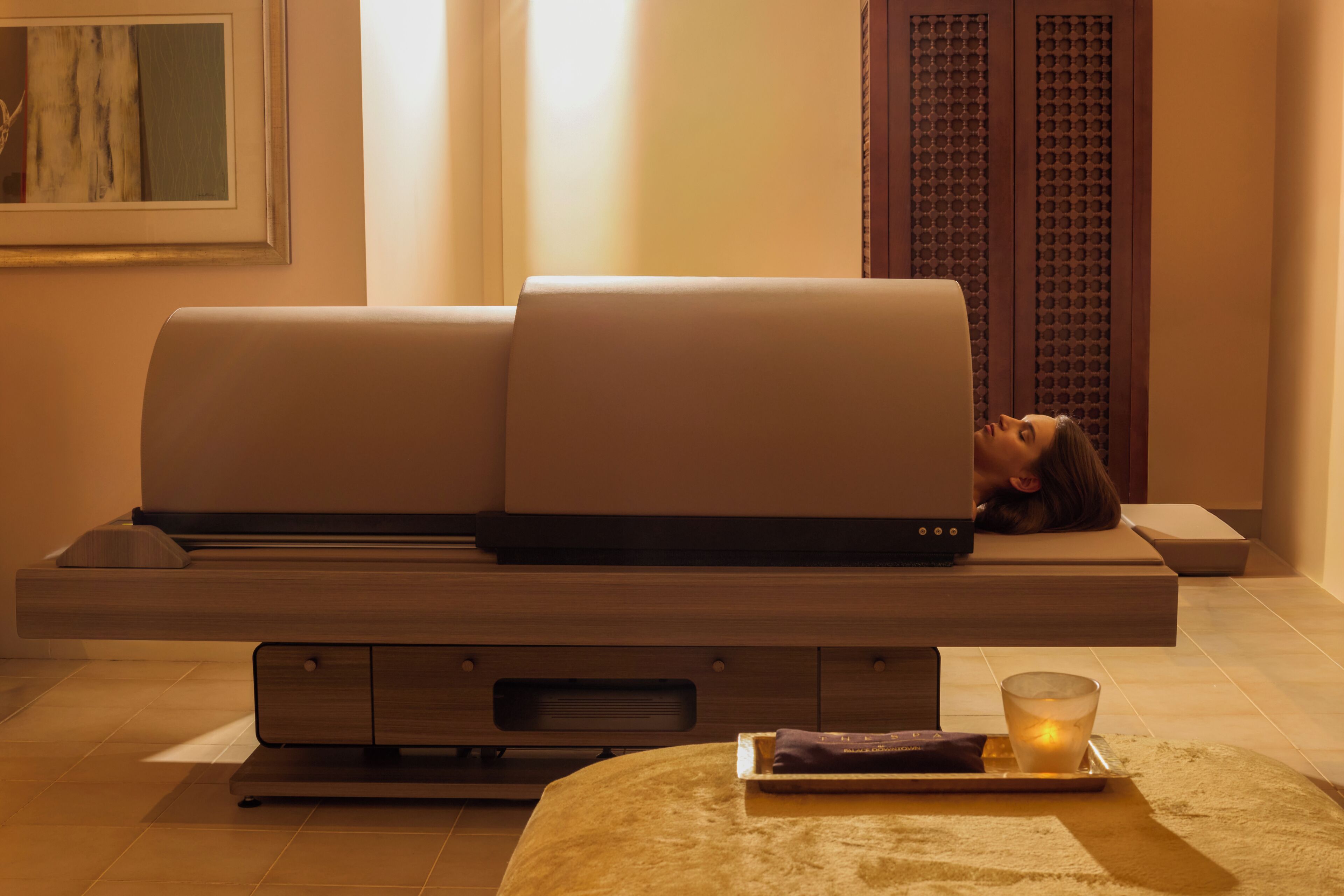 Une table de massage moderne sur laquelle est allongée une personne, une petite table avec une bougie et un haut-parleur fixé au mur.
