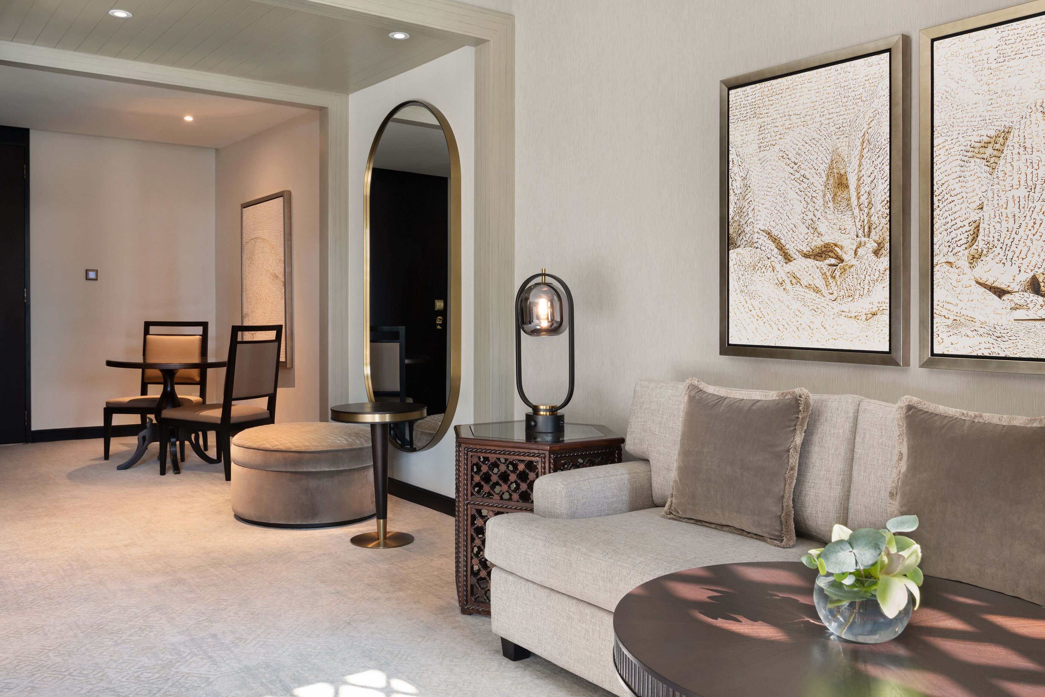A modern living room with a sofa, ottoman, and a round coffee table with a plant. There is a dining area with a table and chairs, and a decorative mirror.