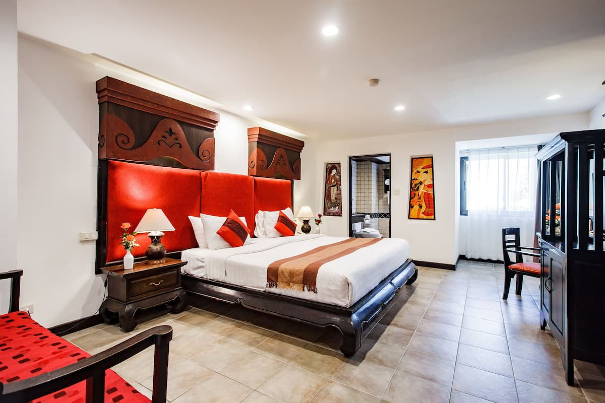 A hotel room with a large bed, a nightstand with a lamp, a chair, and a desk with a red tablecloth.