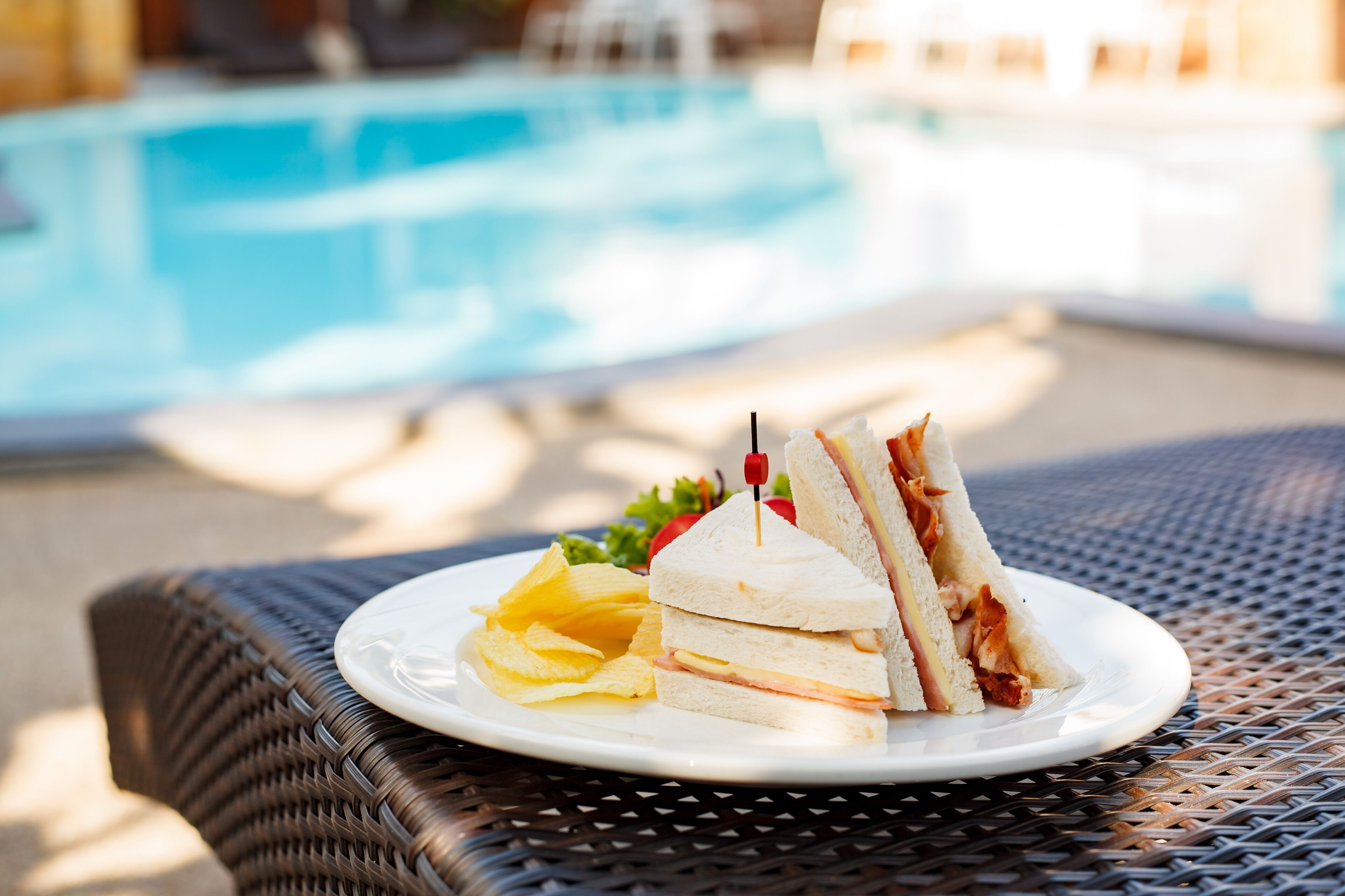 A plate with a sandwich and chips on a wicker table near a pool.