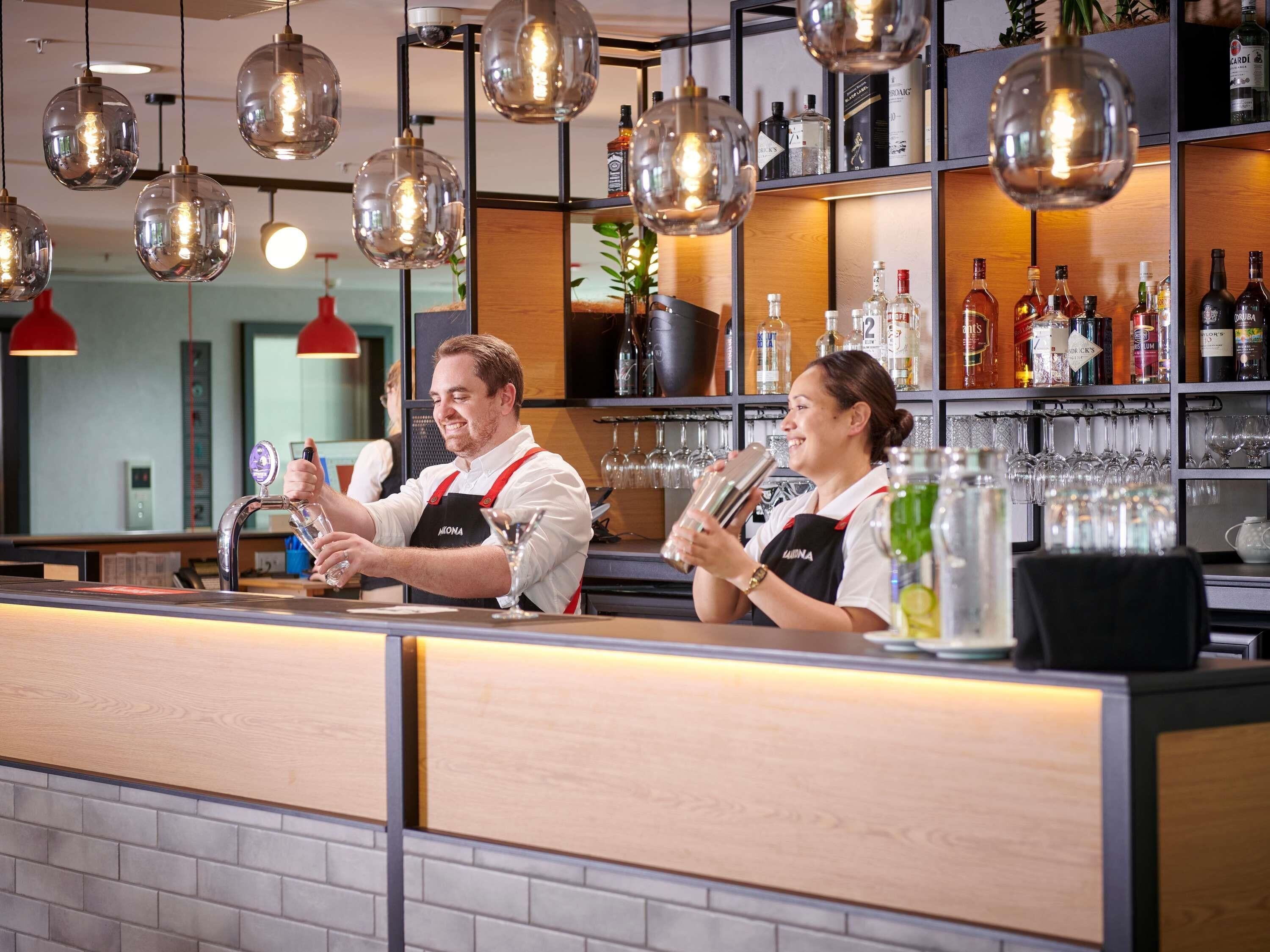 Un espace bar avec des barmen, un comptoir, des luminaires suspendus et des étagères remplies de bouteilles et de verres.