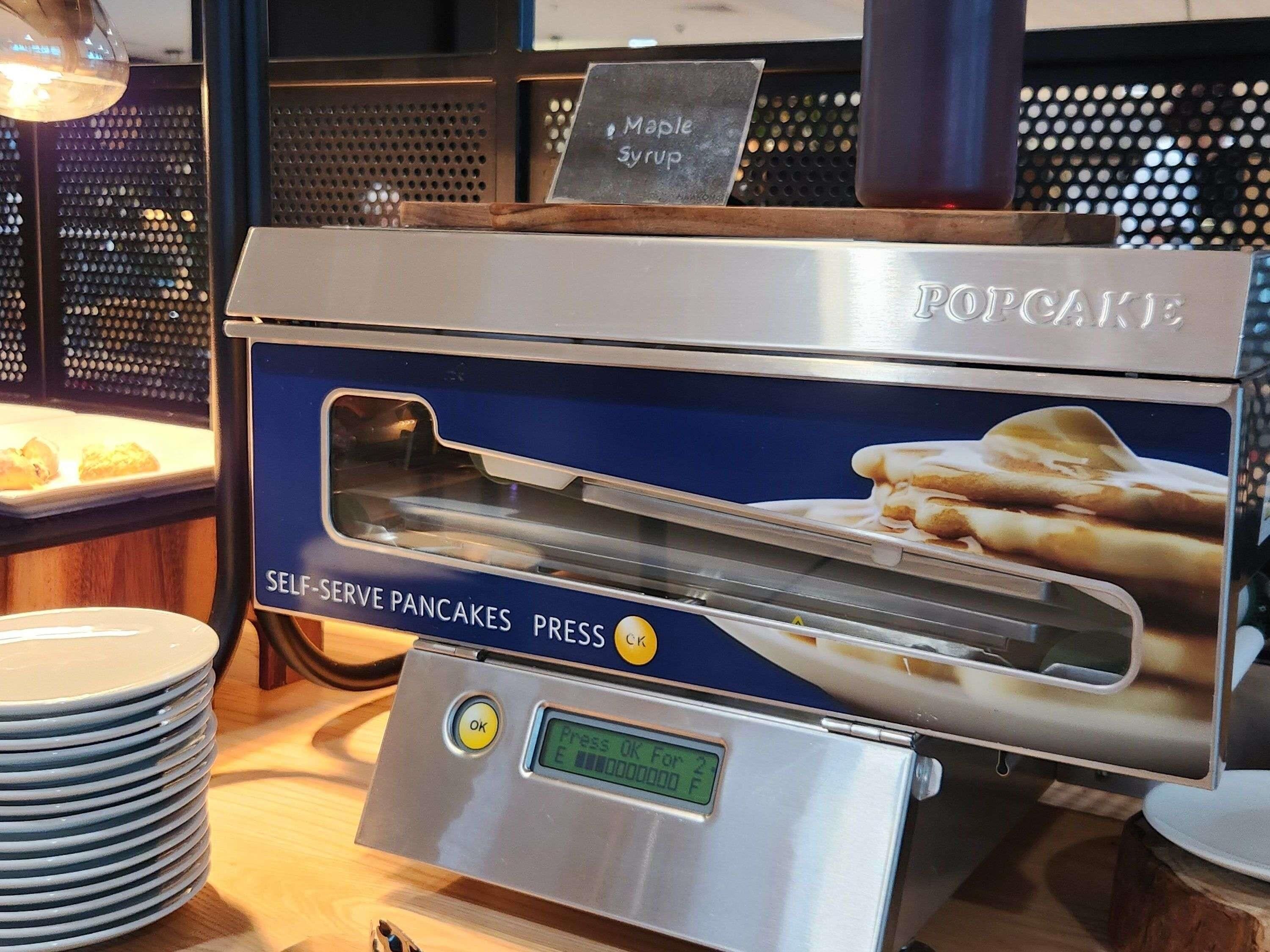 A self-serve pancake press machine with a digital display, a sign for maple syrup, and a stack of plates.