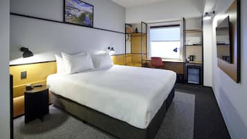 Ein Hotelzimmer mit einem großen Bett, einem Schreibtisch, einem Stuhl, einer kleinen Küchenzeile und einem Fenster mit Blick auf eine Stadtlandschaft.