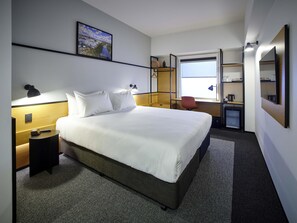 Une chambre d’hôtel avec un grand lit, un bureau, une chaise, une petite kitchenette et une fenêtre donnant sur un paysage urbain.