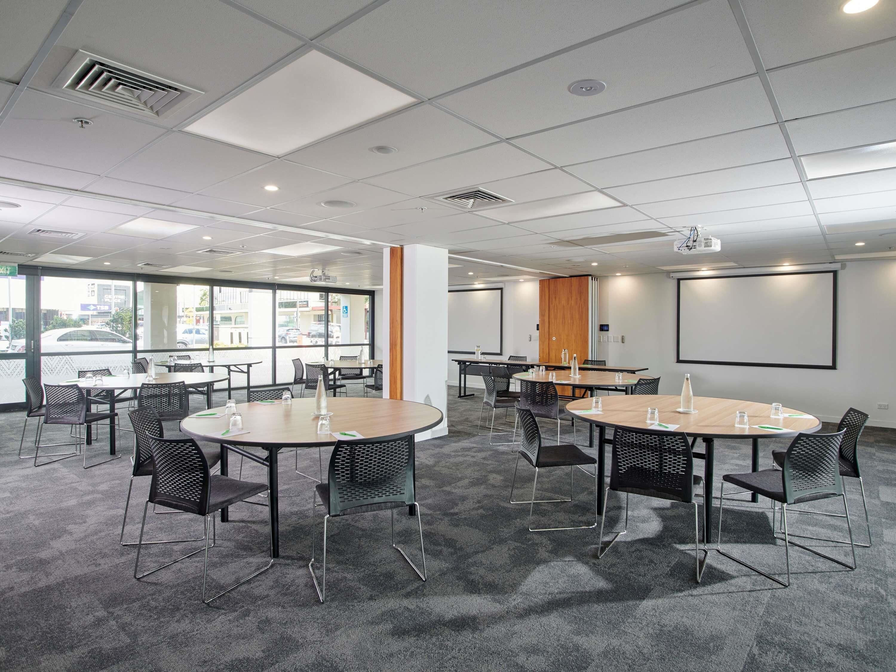 A conference room with round tables, chairs, a projector screen, and a large window.