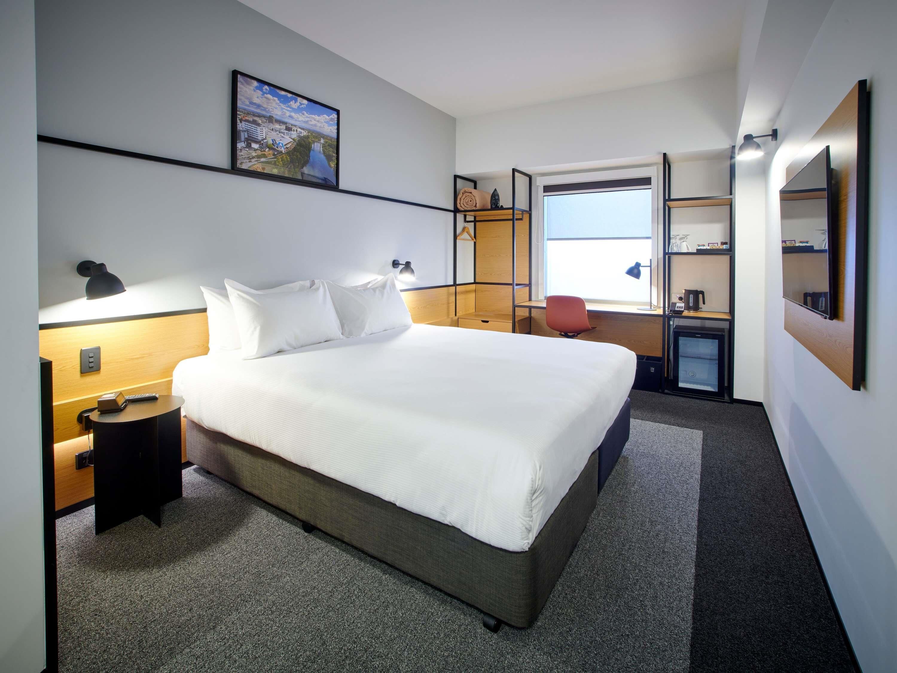 A hotel room with a bed, bedside tables, a desk, a chair, a small kitchenette, a window, and a wall-mounted TV.