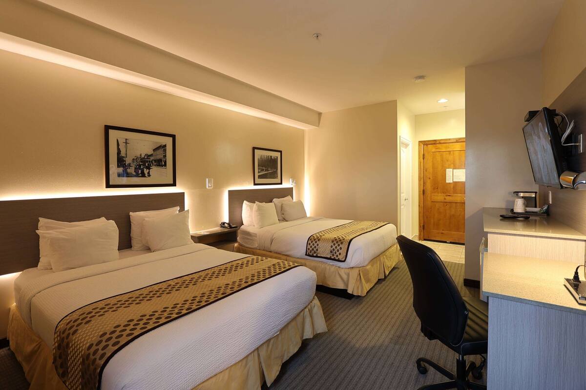 Une chambre d’hôtel avec deux lits, un bureau et une chaise. Des tableaux encadrés ornent le mur, et une porte en bois est visible au fond.