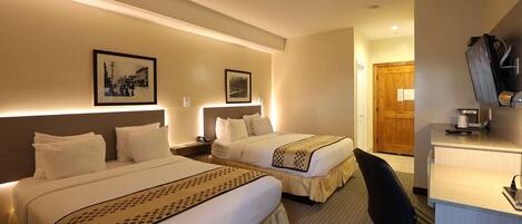 Habitación de hotel con dos camas, un escritorio y una silla. Se observan cuadros en la pared y una puerta de madera al fondo.